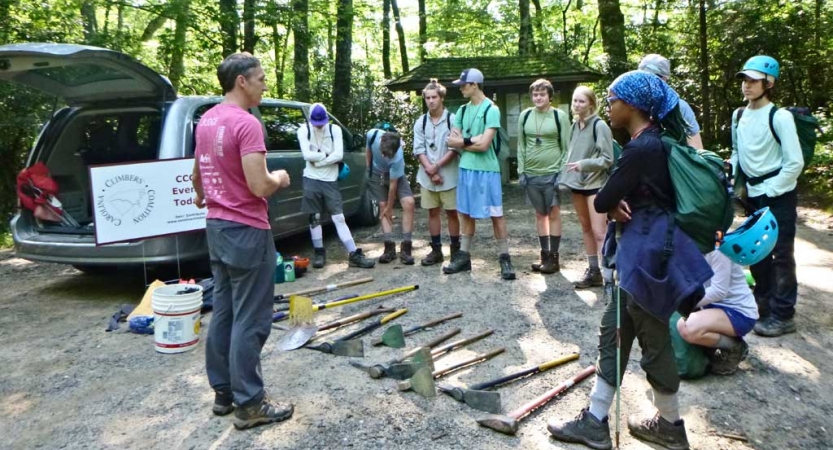 An instructor speaks to a group of students. A selection of gardening tools lay on the ground between them.